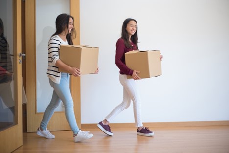 Two women carrying moving boxes into a room.